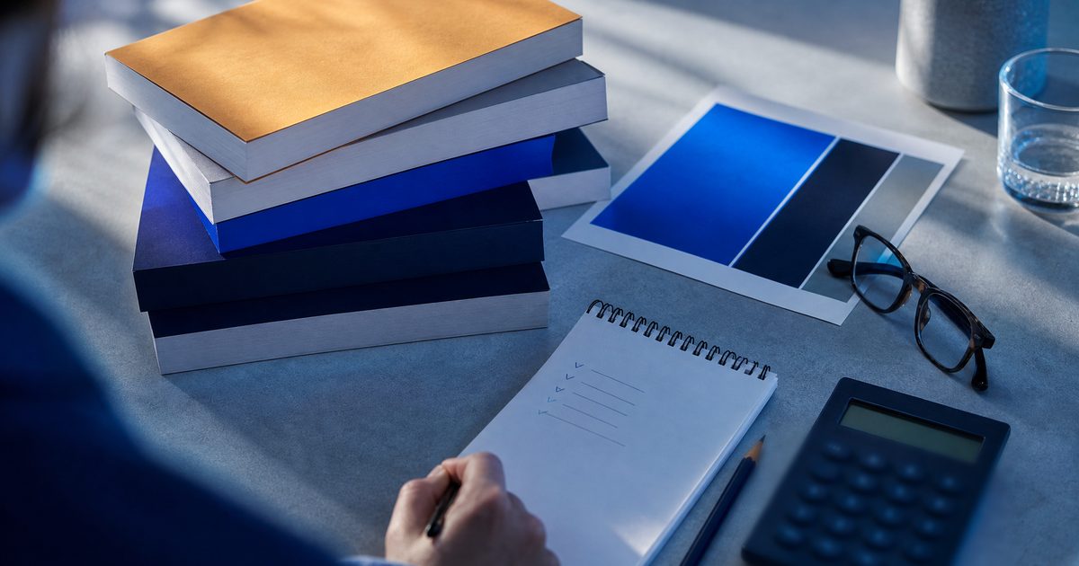 A desk with a stack of paperback proof copies, a handwritten budget on a notepad and a calculator