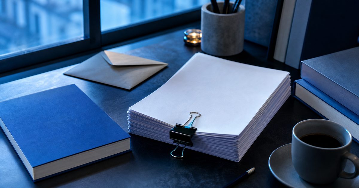 A printed manuscript held by a clip next to a finished paperback on a desk in daylight